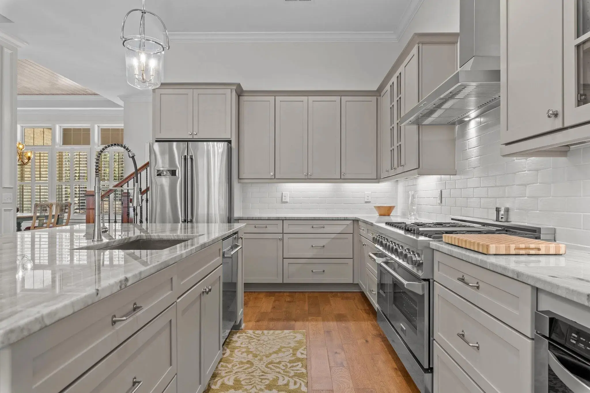 Contemporary kitchen with light gray cabinetry, stainless steel appliances, a large marble island with an undermount sink, and a glossy white subway tile backsplash, complemented by hardwood flooring and a pendant light fixture.