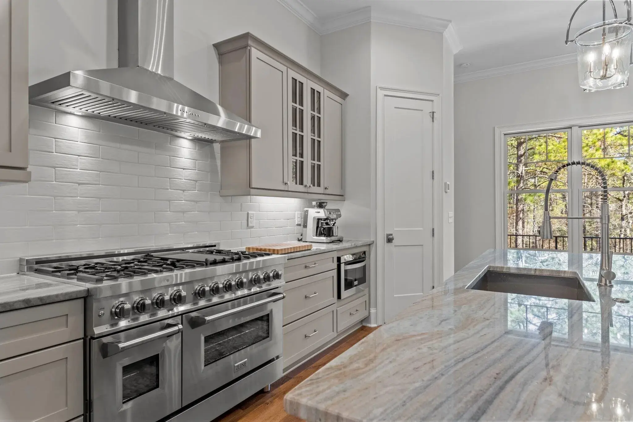 Modern kitchen with stainless steel appliances, including a professional-grade range and hood, light gray cabinetry, and a glossy white subway tile backsplash, complemented by a large marble island with an undermount sink and gooseneck faucet.