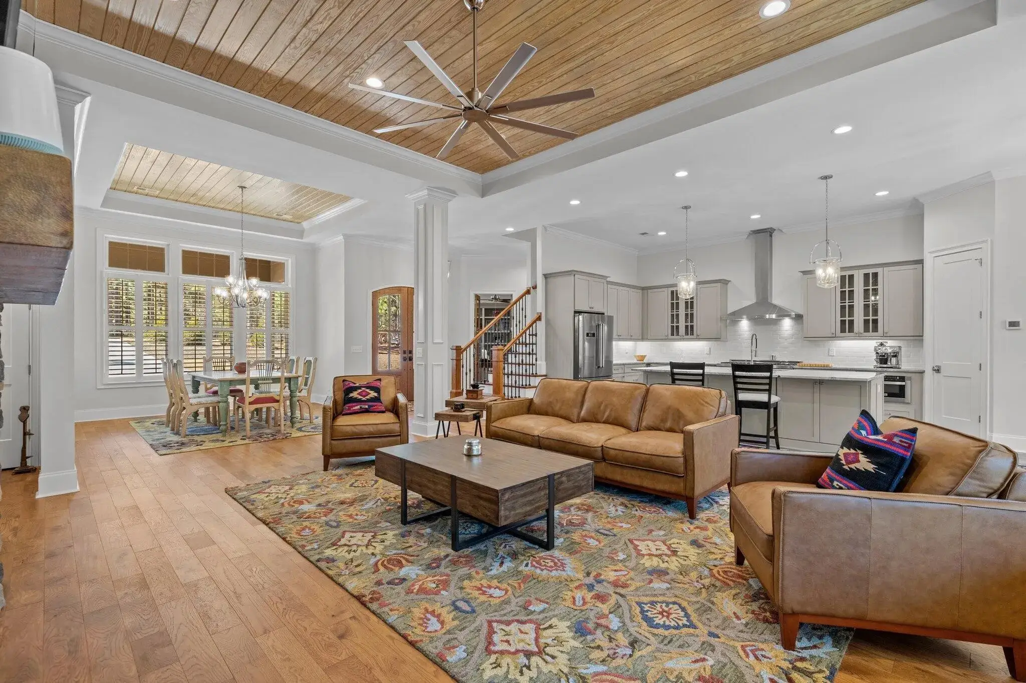 Spacious open-concept living area featuring a cozy seating arrangement with leather sofas, colorful area rugs, and a wooden coffee table, adjacent to a modern kitchen with light gray cabinetry and a dining area under a tray ceiling with a wooden finish.