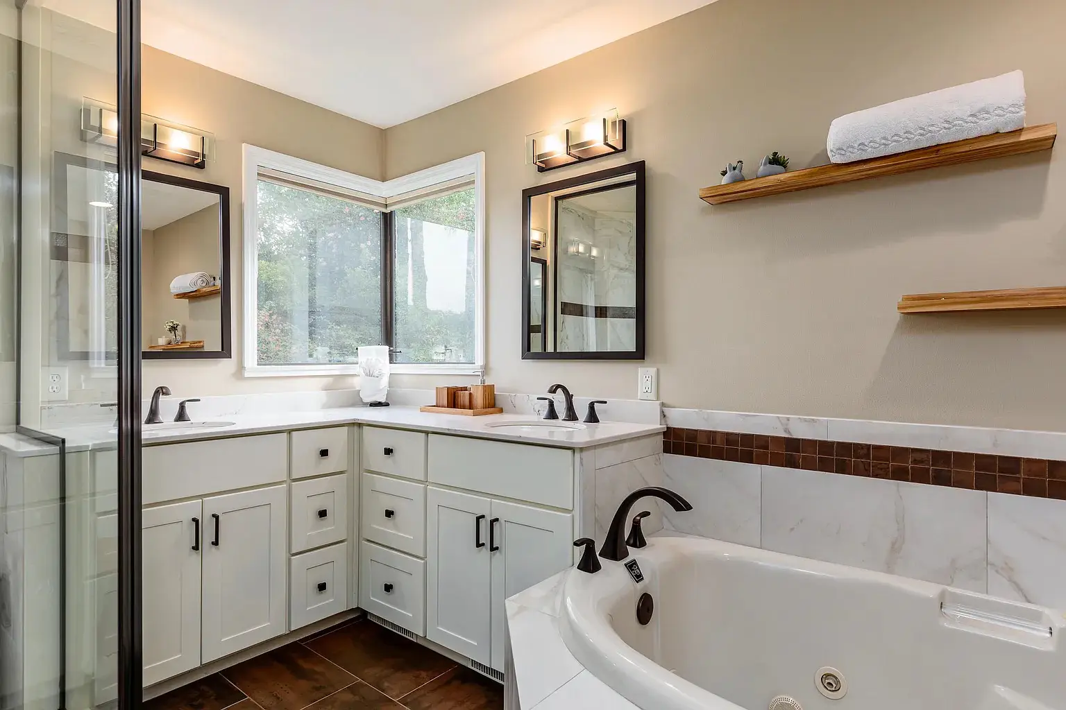 Cozy bathroom featuring a corner vanity with white cabinetry and black fixtures, a large soaking tub with a tiled surround, open wooden shelves for storage, and a glass-enclosed shower, all illuminated by natural light from large windows.