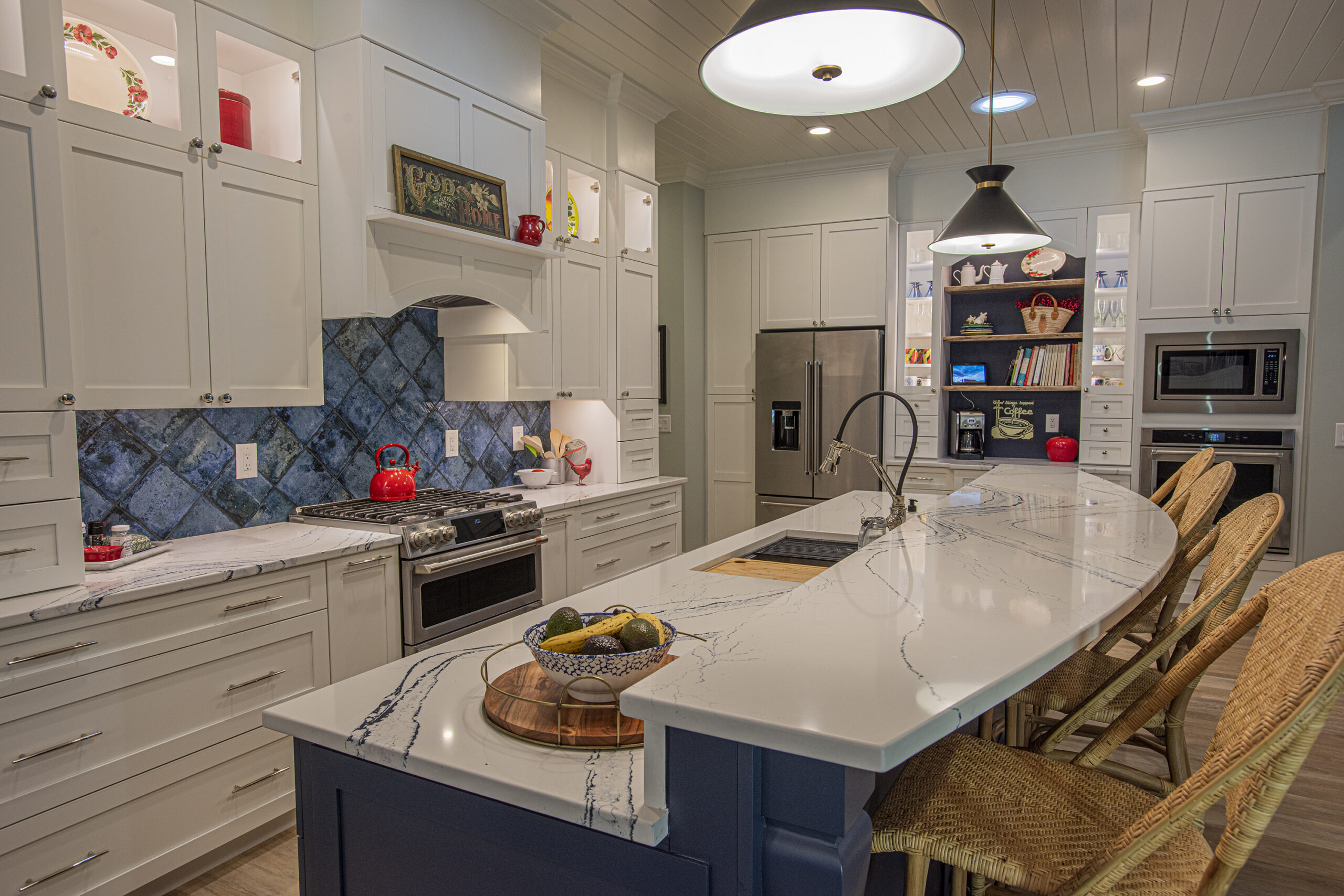 Modern kitchen with a large white marble island, wicker bar stools, and stainless steel appliances. The kitchen features white cabinetry with glass display shelves, a blue tile backsplash, and a decorative sign above the stove. The space is warmly lit with pendant lights, creating a cozy and inviting atmosphere.