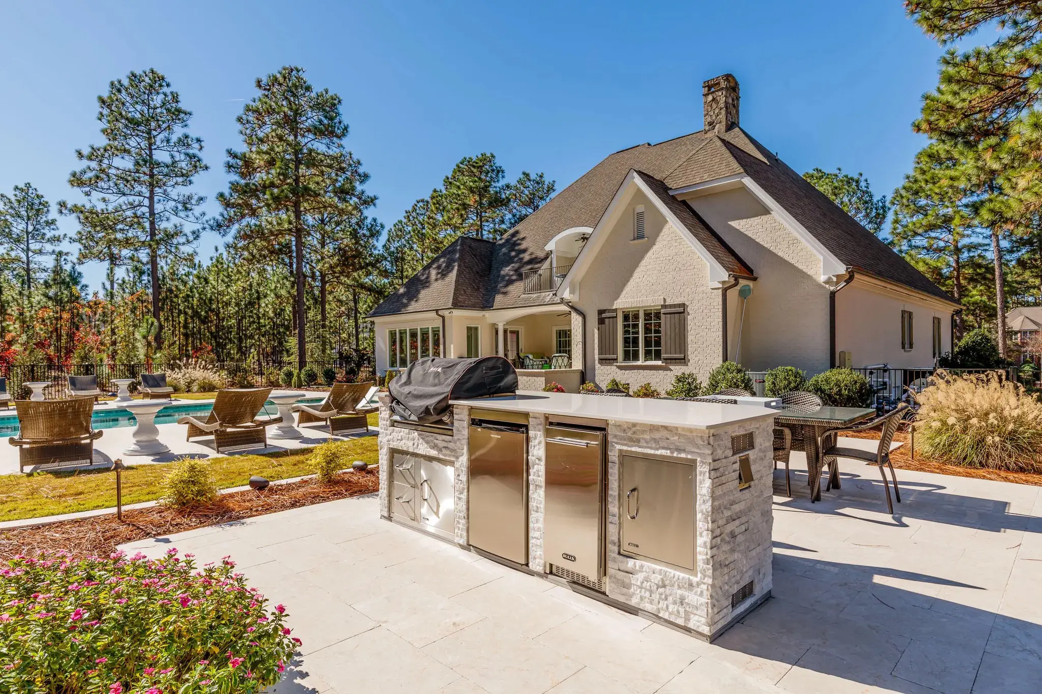 Beautiful outdoor patio area with a built-in grill and stainless steel appliances, overlooking a swimming pool with lounge chairs, surrounded by lush greenery and tall pine trees, adjacent to a charming brick house with a steep roof.