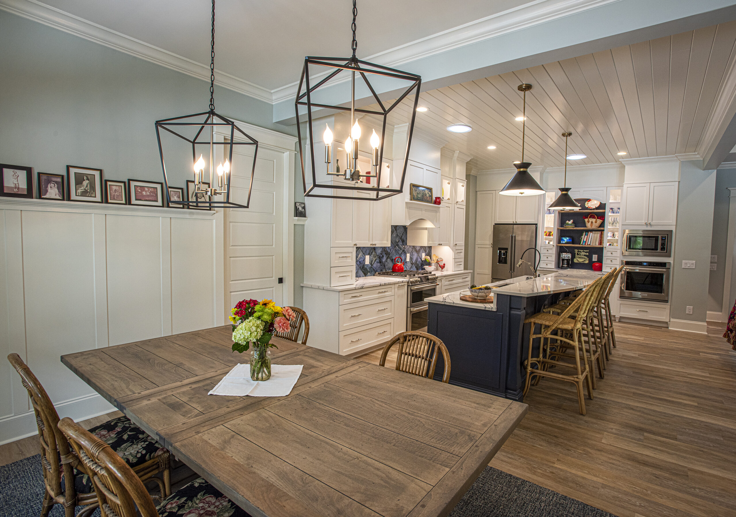 Spacious kitchen and dining area featuring a rustic wooden dining table with a vase of flowers, wicker chairs, and modern pendant lights. The kitchen showcases white cabinetry, a large island with bar stools, and stainless steel appliances, complemented by a wainscoted wall adorned with framed photographs.