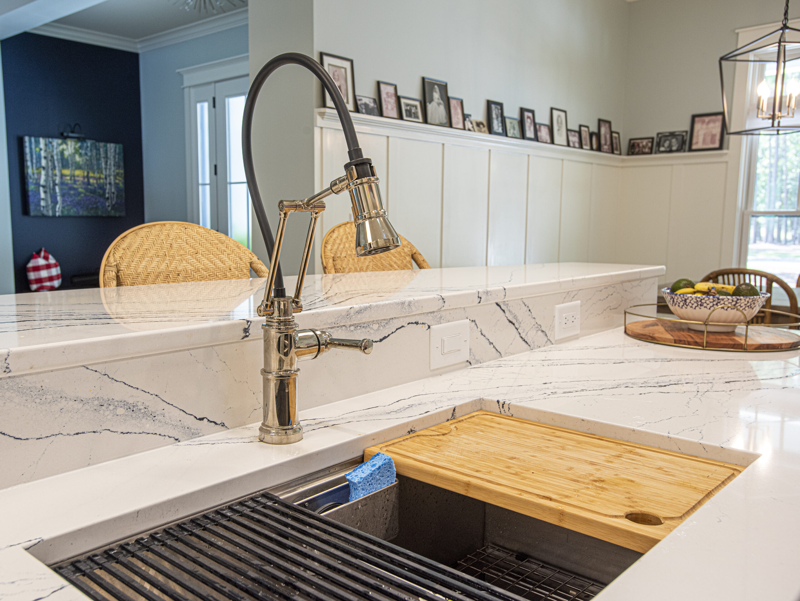 Close-up of a modern kitchen sink with a sleek pull-down faucet, integrated cutting board, and marble countertops, with a glimpse of wicker bar stools and framed photos along a wainscoted wall in the background.