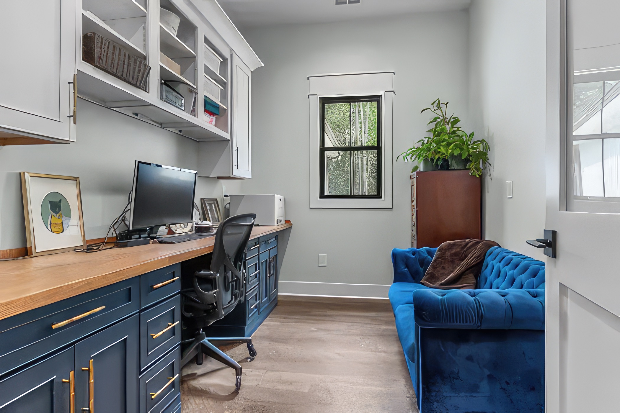 Charming home office featuring navy blue cabinetry with brass hardware, a wooden countertop, open shelving, a cozy blue tufted sofa, and a single window letting in natural light, complemented by light walls and wood flooring.