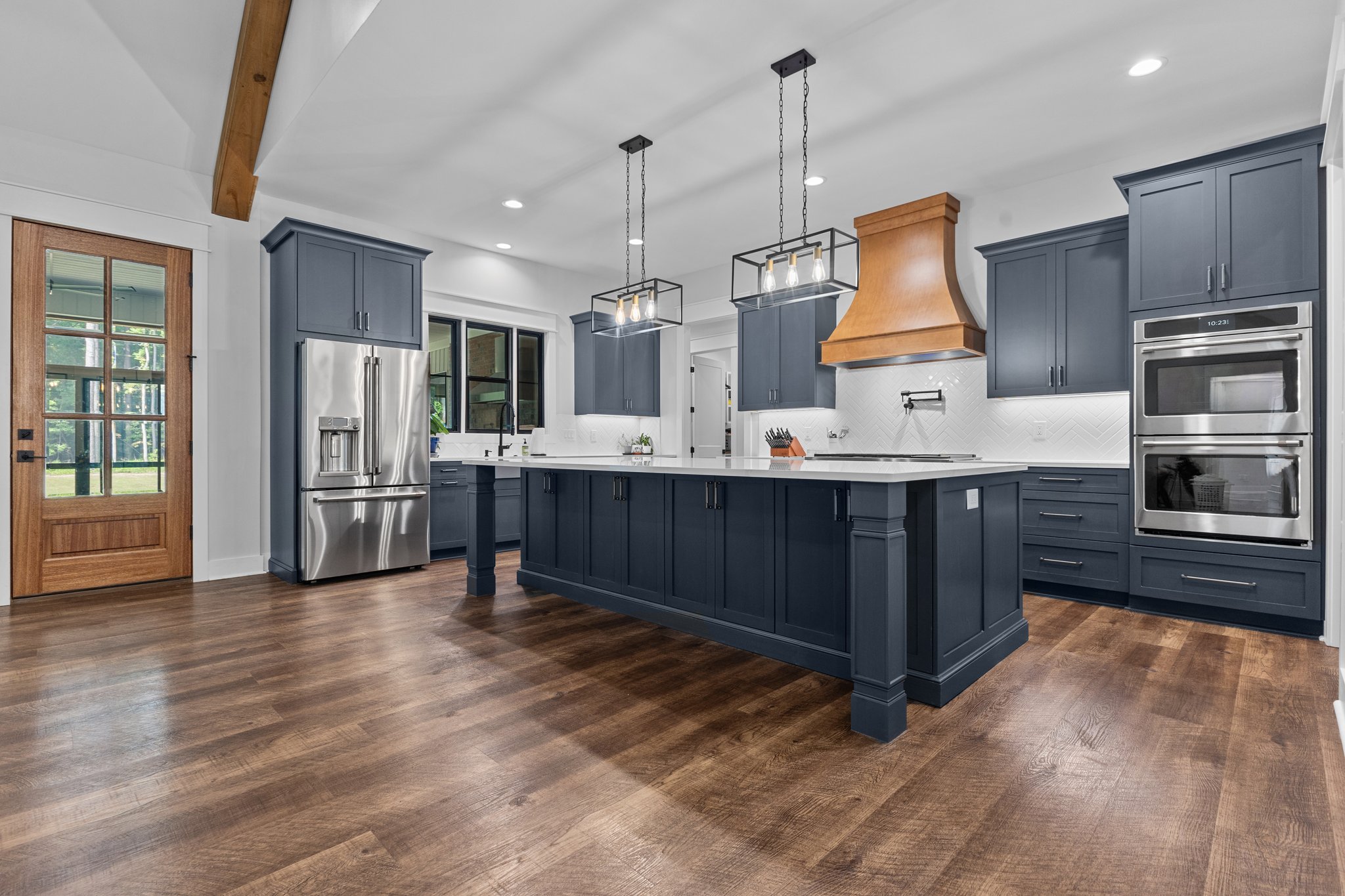 Modern kitchen with dark blue cabinetry, a large white quartz island, and stainless steel appliances, including a double oven and a refrigerator. The kitchen features a wood vent hood over the stove, a white herringbone tile backsplash, and two black metal pendant lights. The space is accented with wood flooring and a natural wood door with glass panels leading to the outside.