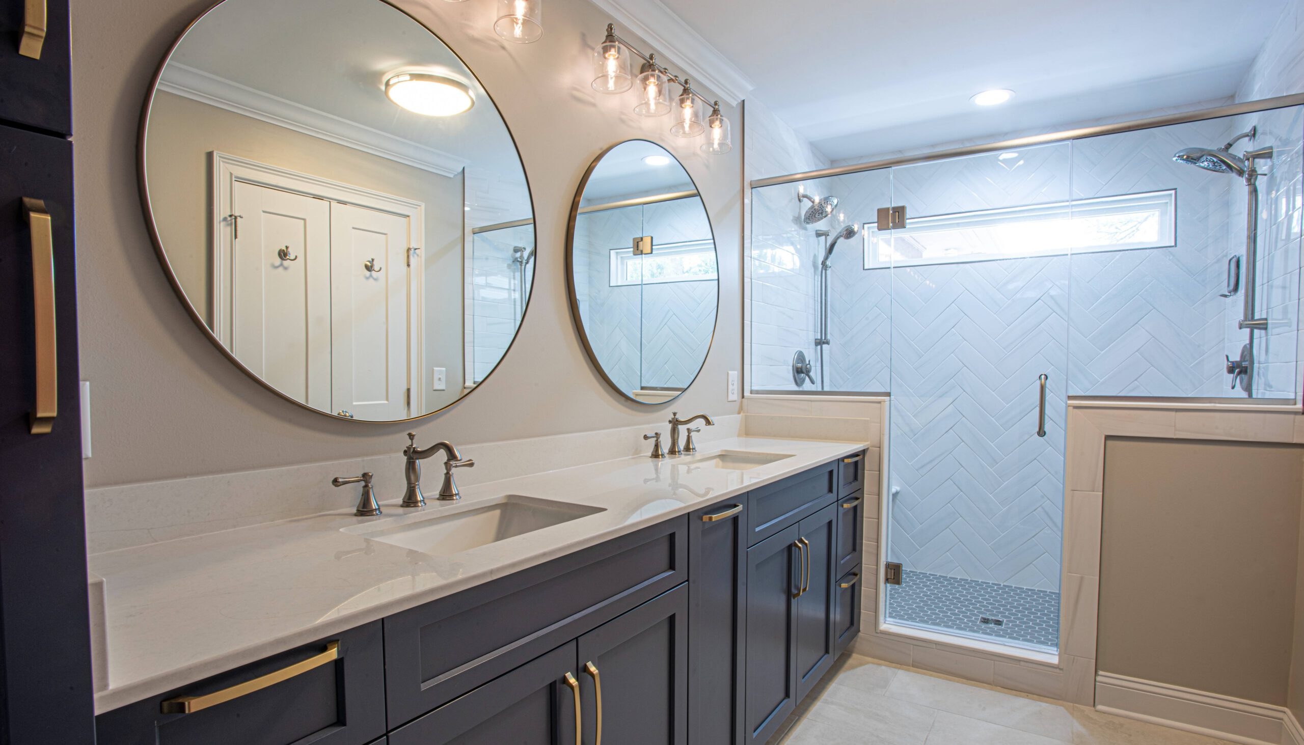 Modern bathroom with a double vanity featuring navy blue cabinetry, white quartz countertops, and round mirrors, complemented by brushed nickel fixtures and glass pendant lights, adjacent to a glass-enclosed shower with herringbone tile walls.