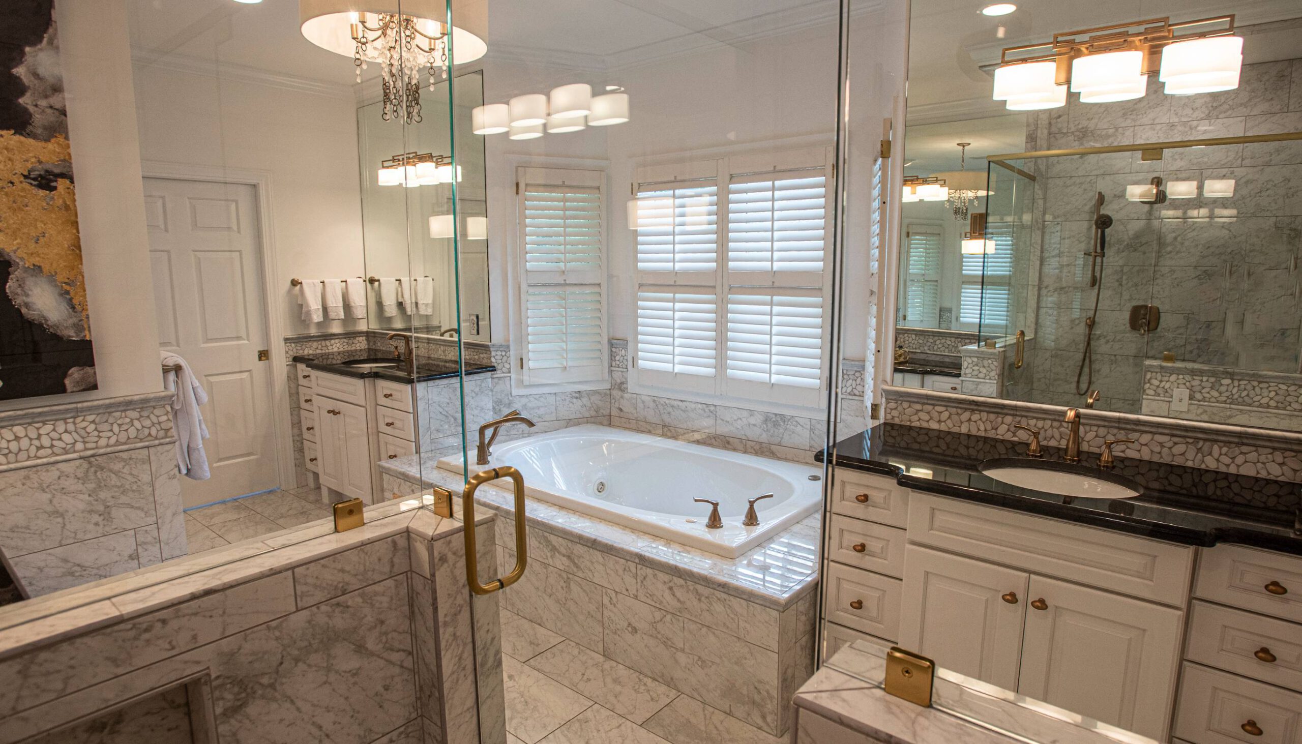 Luxurious bathroom with a large soaking tub surrounded by marble tile, glass-enclosed shower, dual vanities with black countertops, gold fixtures, and elegant lighting, accented by white plantation shutters and a chandelier.