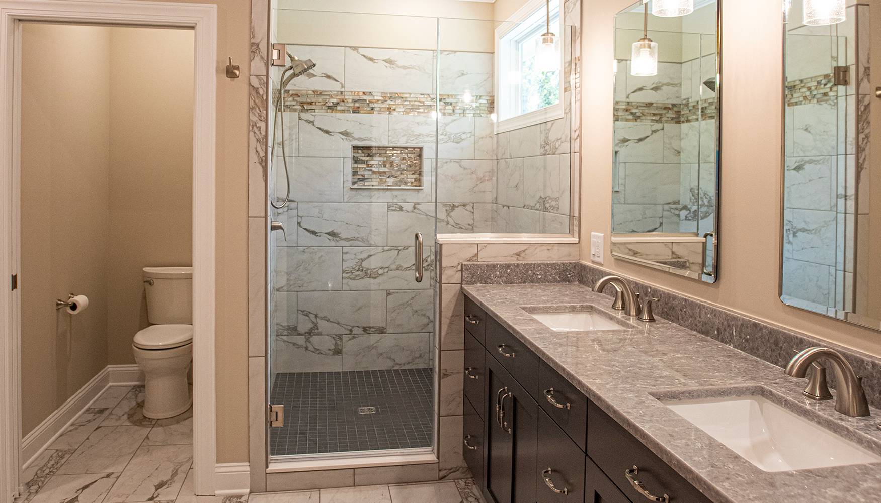 Luxurious bathroom with a glass-enclosed shower featuring marble tile and mosaic accent, double vanity with gray granite countertops, and a separate toilet area with matching marble flooring.