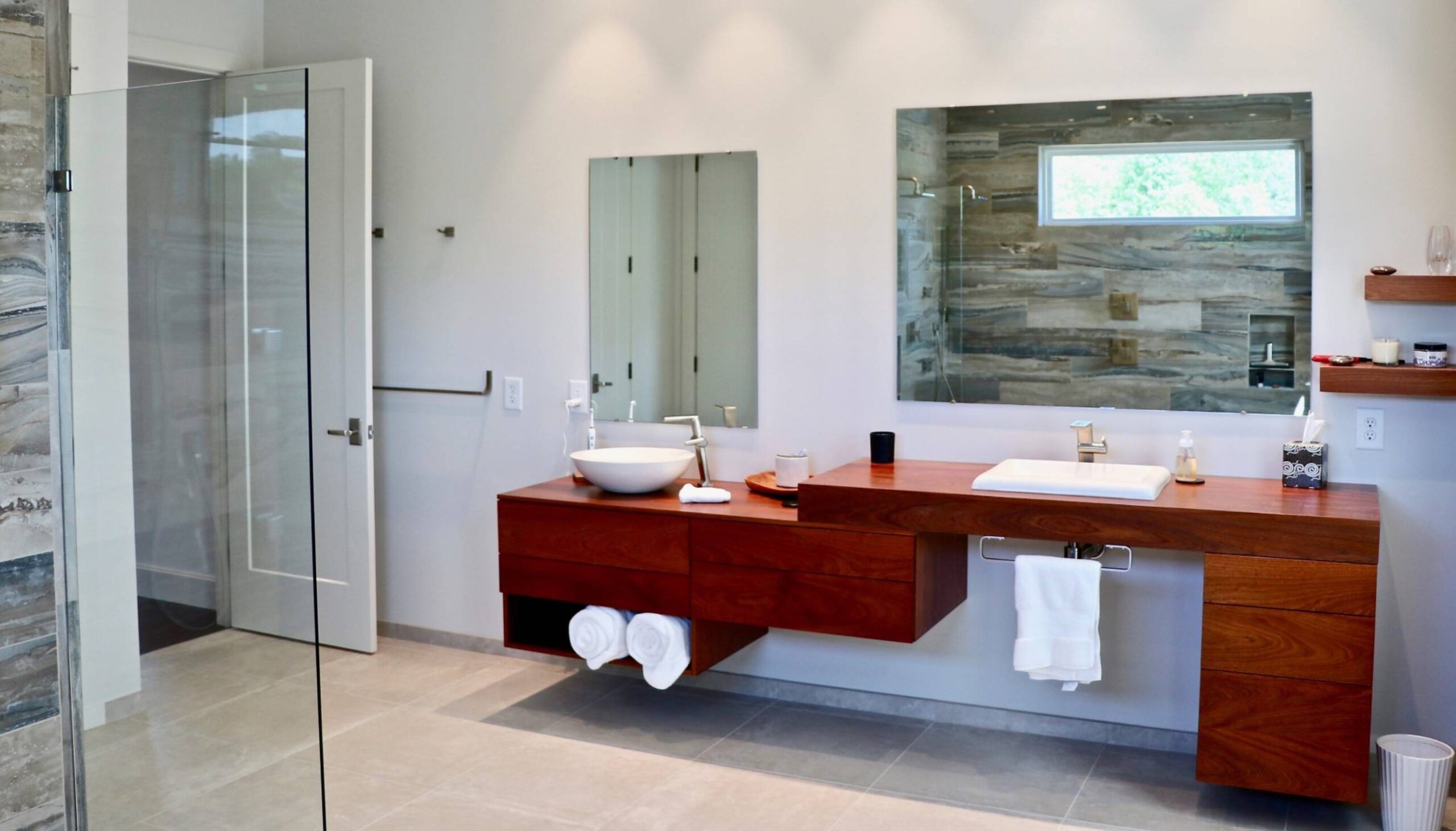 Contemporary bathroom with floating wooden vanities, a vessel sink on one side, and an integrated sink on the other, complemented by large mirrors and open shelving, adjacent to a glass-enclosed shower with stone-textured tile walls.