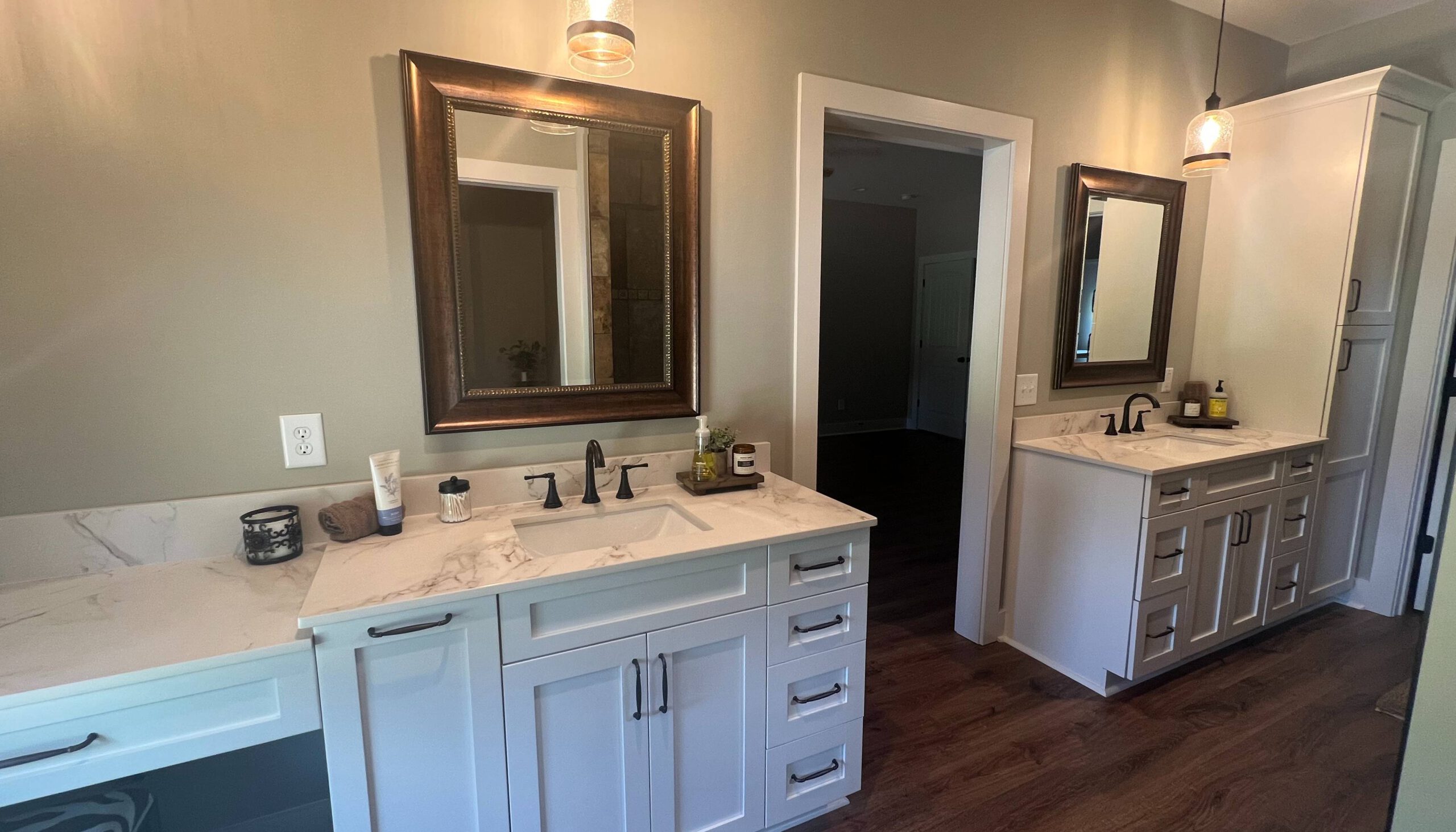 Spacious bathroom with dual vanities featuring white cabinetry, marble countertops, and framed mirrors, complemented by dark bronze fixtures and pendant lighting, set against warm wood flooring.