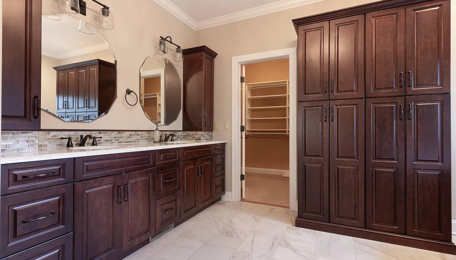 Traditional bathroom with rich dark wood cabinetry, a double vanity with marble countertops, octagonal mirrors, and glass pendant lighting, adjacent to a spacious walk-in closet, all set on polished marble flooring.