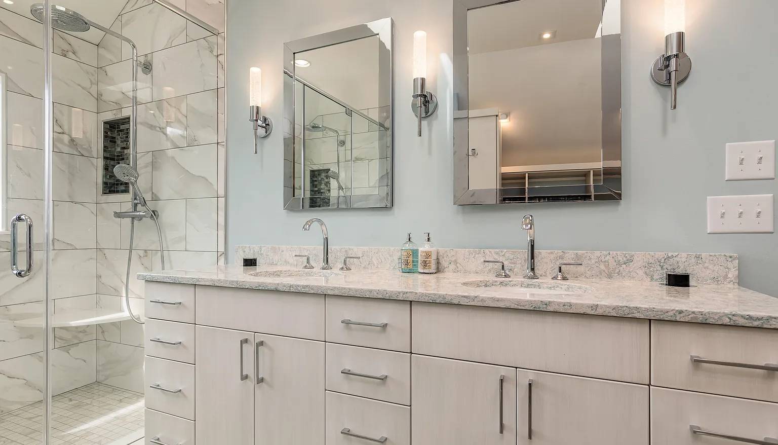 Modern bathroom featuring a double vanity with light wood cabinetry, marble countertops, and chrome fixtures, paired with frameless mirrors and sleek wall sconces, adjacent to a glass-enclosed shower with marble tile and a built-in bench.