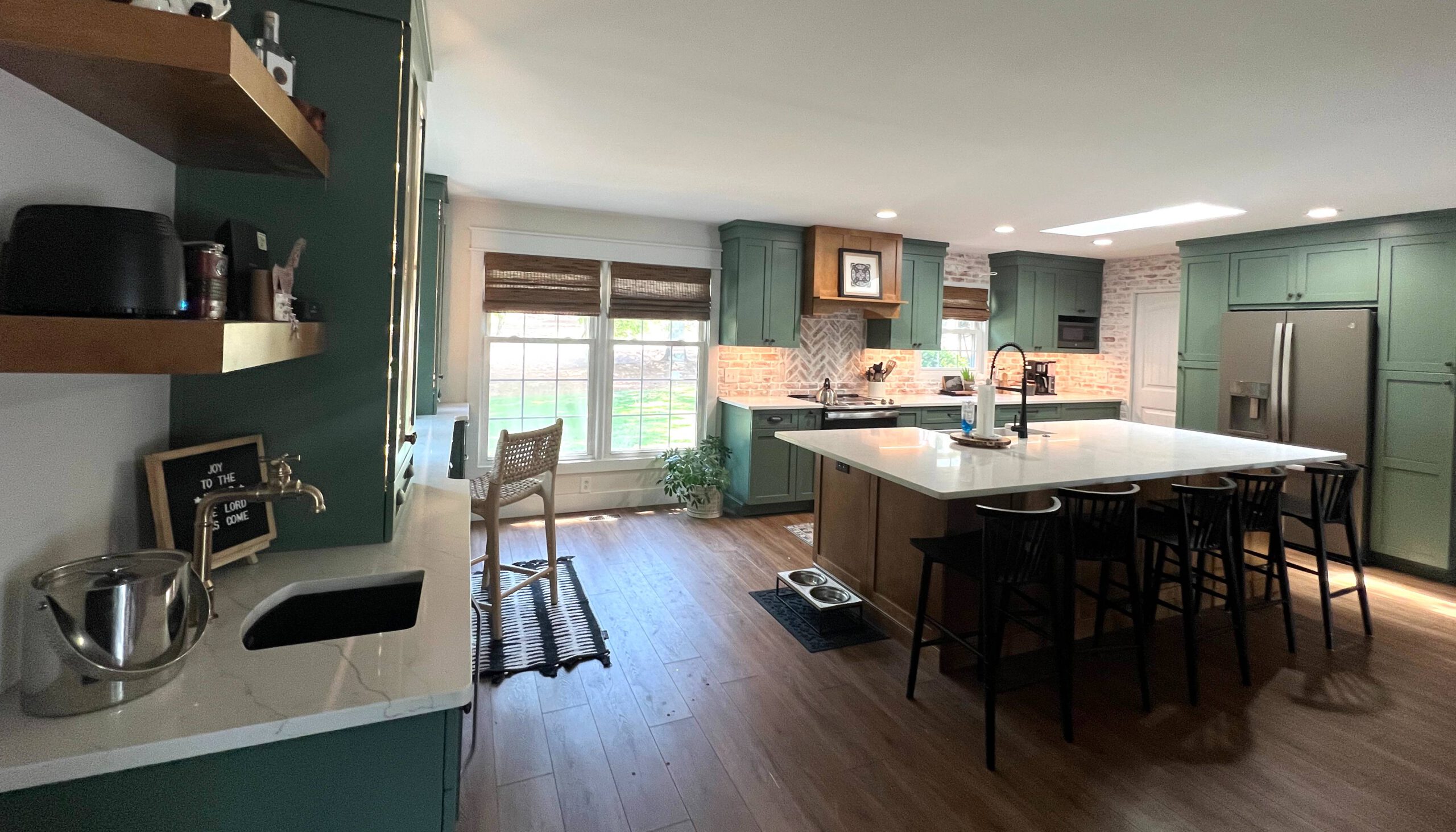 Spacious kitchen with green cabinetry, a large white island with a built-in sink and seating for four, stainless steel appliances, wood flooring, and open shelving, accented by natural light from large windows with woven blinds.