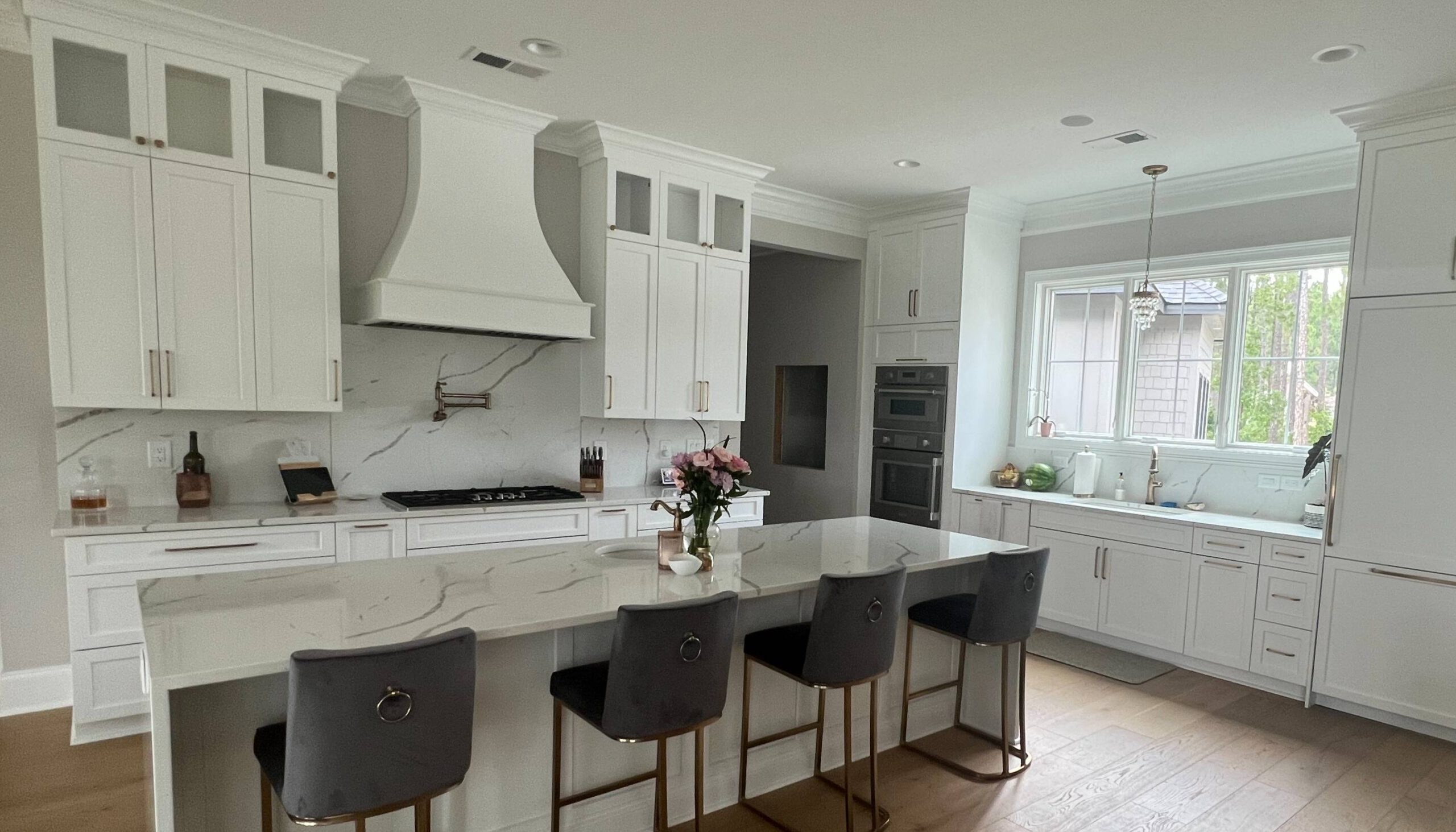 Elegant kitchen with white cabinetry, a large marble island with gray velvet barstools, stainless steel appliances, a custom range hood, and a marble backsplash, accented by natural light from a large window over the sink.