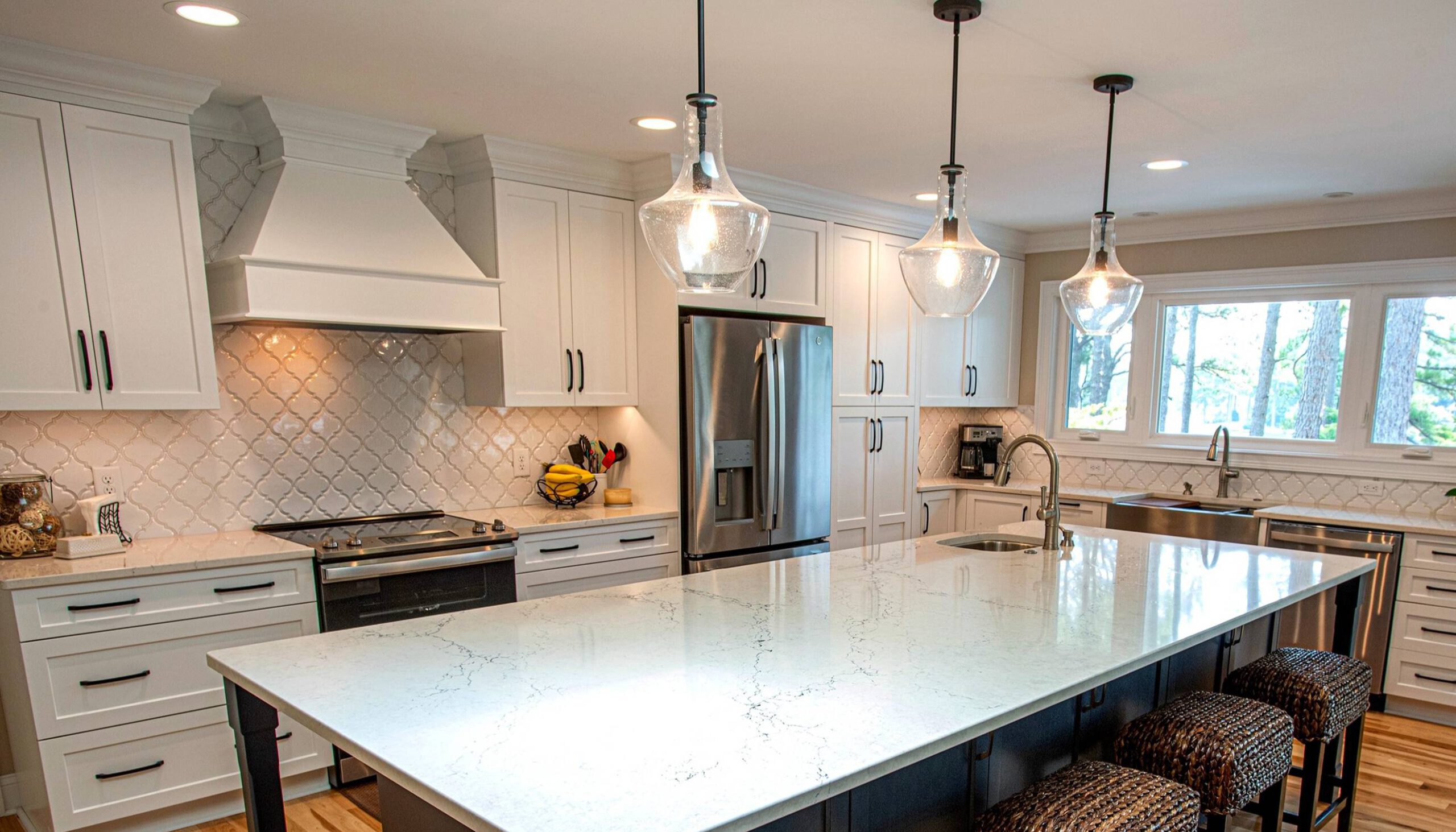 Bright and airy kitchen featuring white cabinetry, a large quartz island with woven barstools, stainless steel appliances, and a patterned tile backsplash, illuminated by glass pendant lights and natural light from large windows.