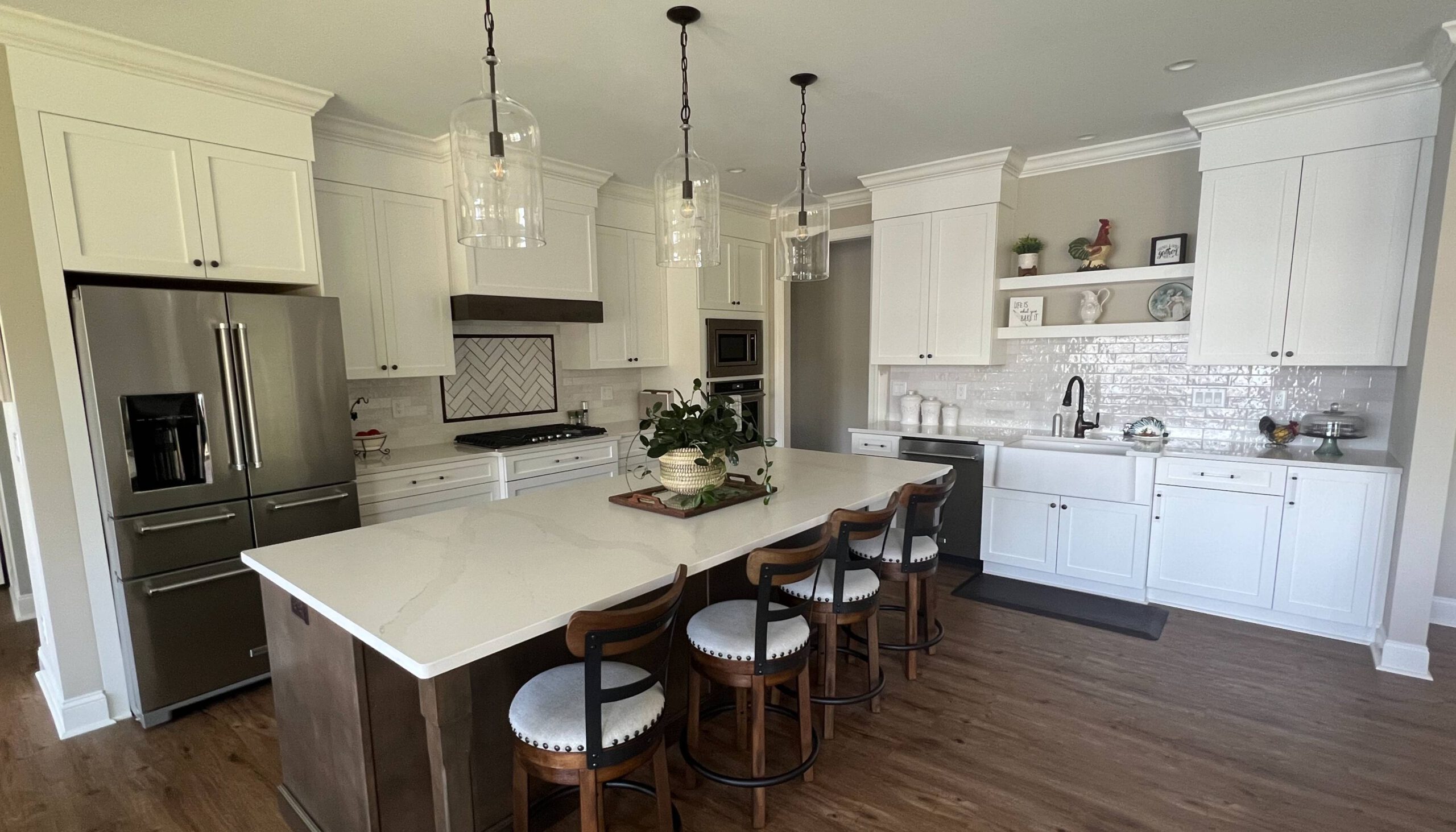 Modern kitchen with white cabinetry, a large quartz island with wooden barstools, stainless steel appliances, and a farmhouse sink, complemented by a herringbone tile backsplash, glass pendant lights, and wood flooring.