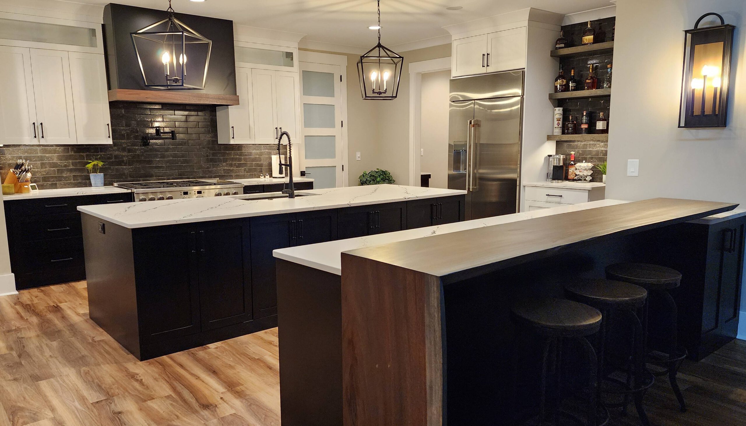 Stylish kitchen with a contrasting black and white color scheme, featuring dark cabinetry, a large island with a marble countertop, stainless steel appliances, a wood-accented range hood, and modern pendant lighting, complemented by a breakfast bar with wooden stools and a warm wood floor.