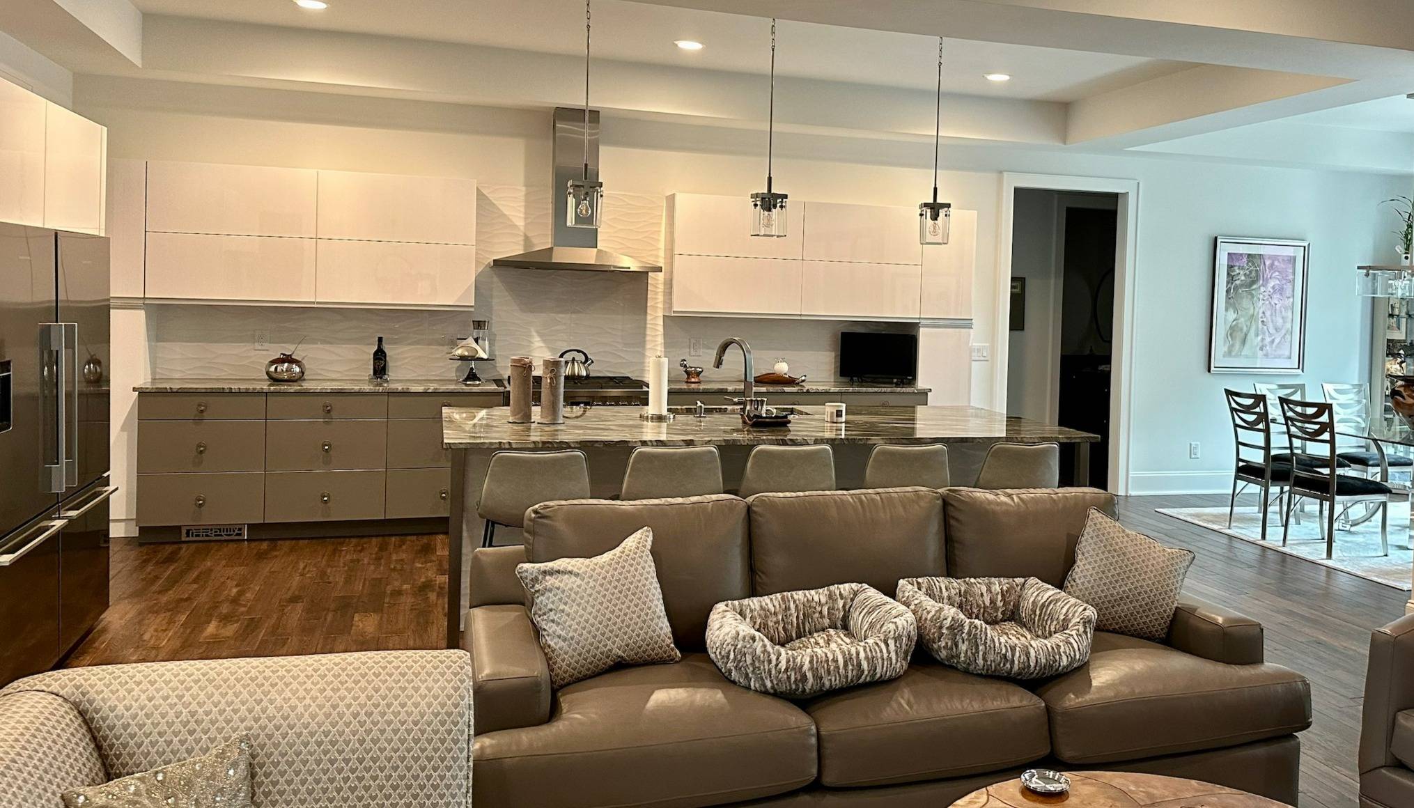 Open-concept kitchen and living area featuring sleek cabinetry, a large island with seating, stainless steel appliances, and modern pendant lighting, with a comfortable seating area in the foreground and a dining area in the background.