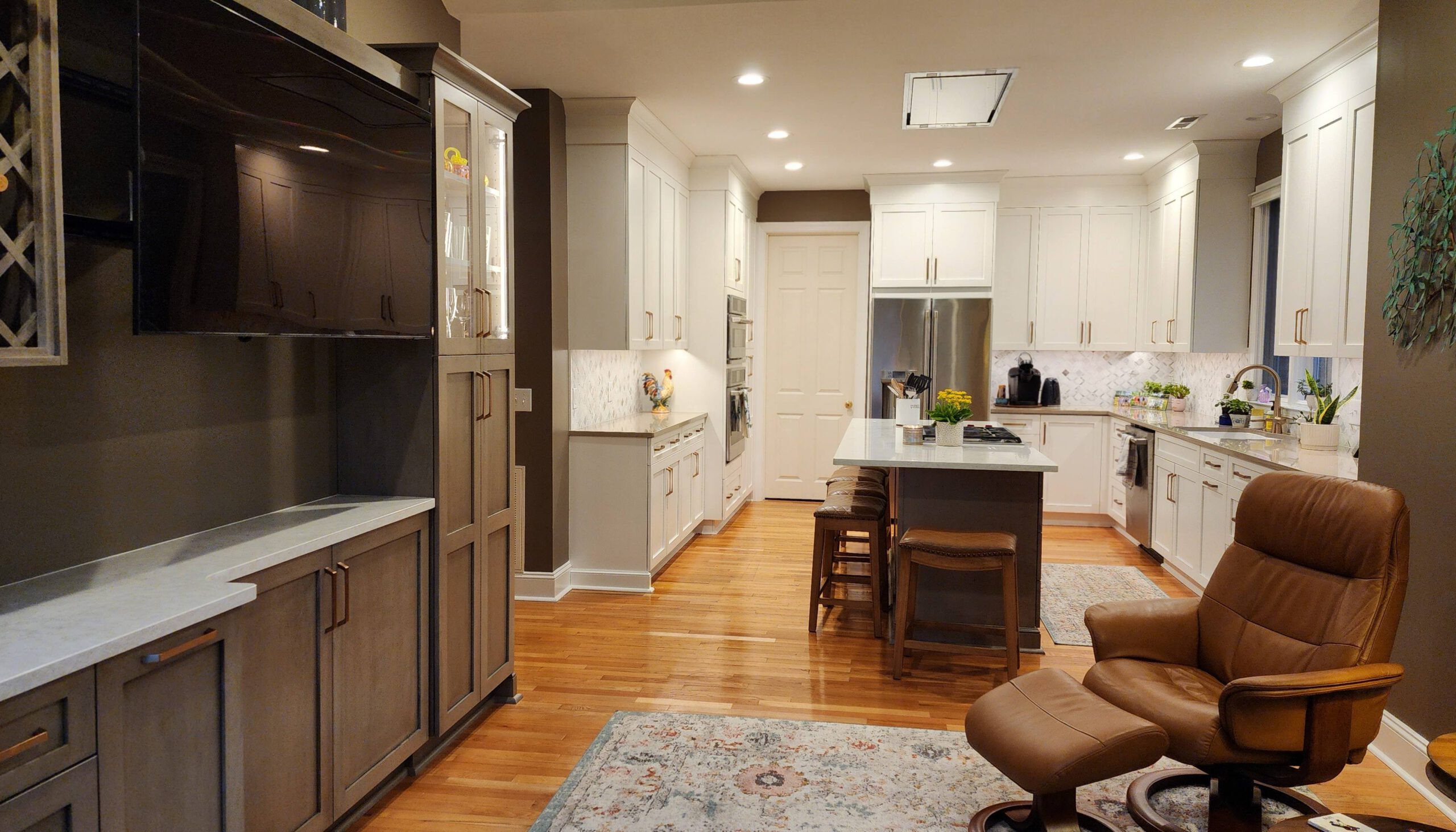 Cozy kitchen and living space featuring white cabinetry, a large island with barstools, stainless steel appliances, and wood flooring, complemented by warm-toned walls, a built-in entertainment center, and a leather recliner with an ottoman.