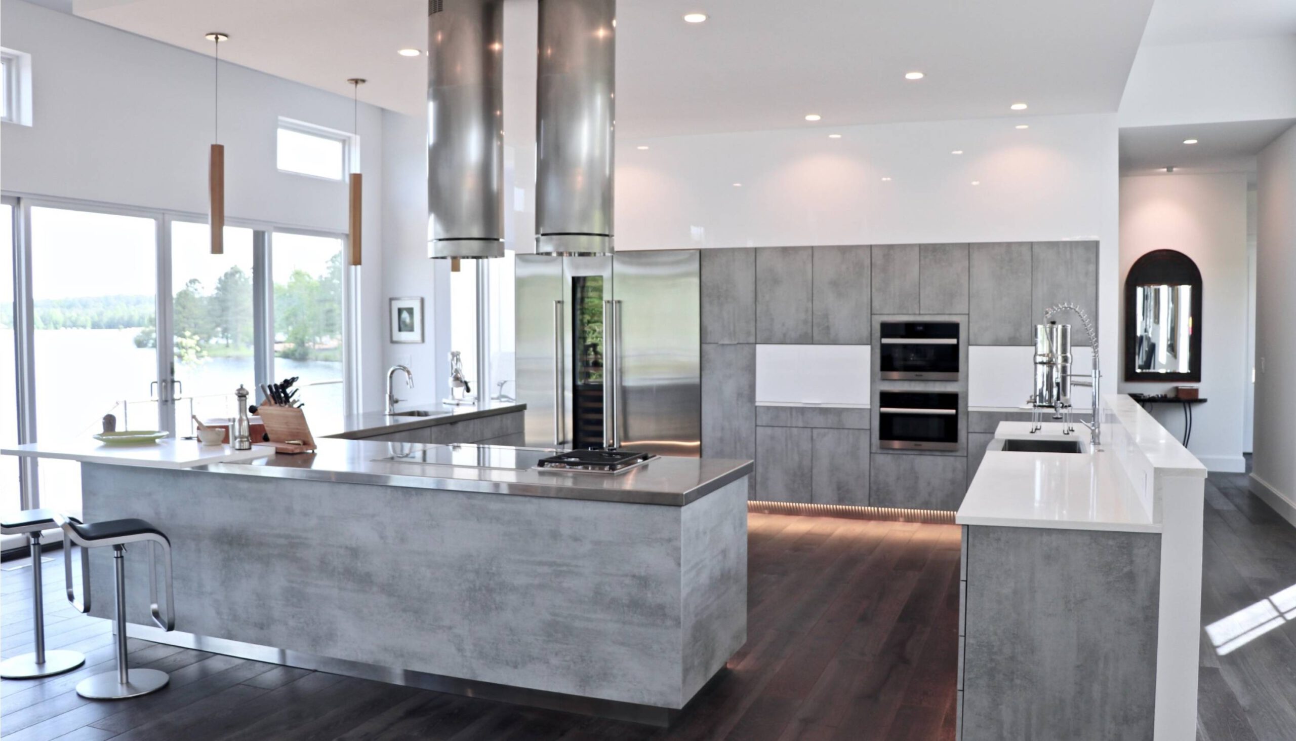 Sleek, modern kitchen featuring gray concrete-style cabinetry and islands, stainless steel appliances, and large pendant lights, with expansive windows offering a view of the outdoors and filling the space with natural light.