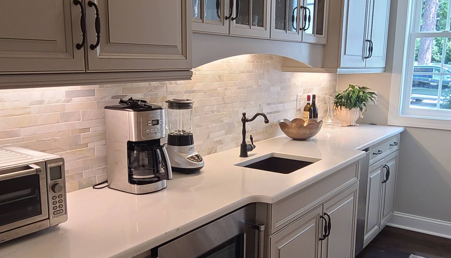 Elegant butler's pantry with white cabinetry, a white quartz countertop, and a neutral-toned tile backsplash, featuring a coffee maker, blender, toaster oven, and a small sink with a bronze faucet, all illuminated by under-cabinet lighting.