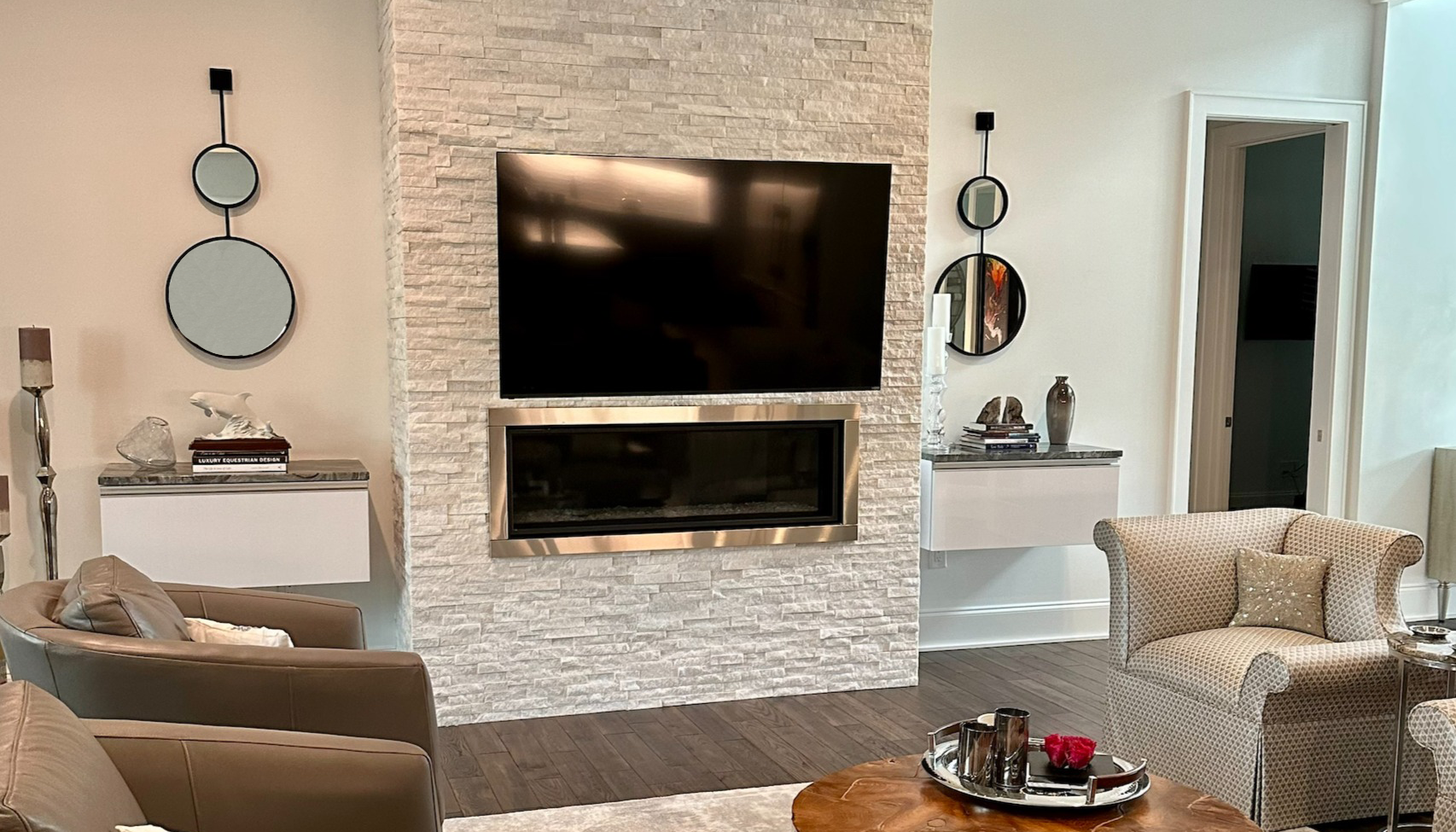 Contemporary living room featuring a wall-mounted flat-screen TV and a modern linear fireplace set in a white stacked stone wall, flanked by floating cabinets with decorative items, round mirrors, and accent chairs on dark wood flooring.